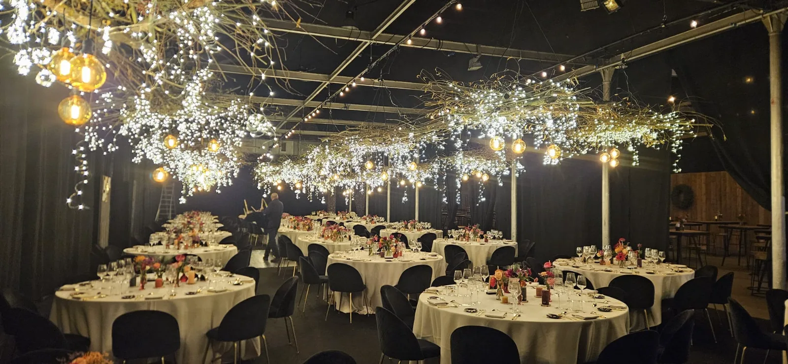 Feestzaal met ronde gedekte tafels onder hangende takken vol lichtjes en amberkleurige bollampen