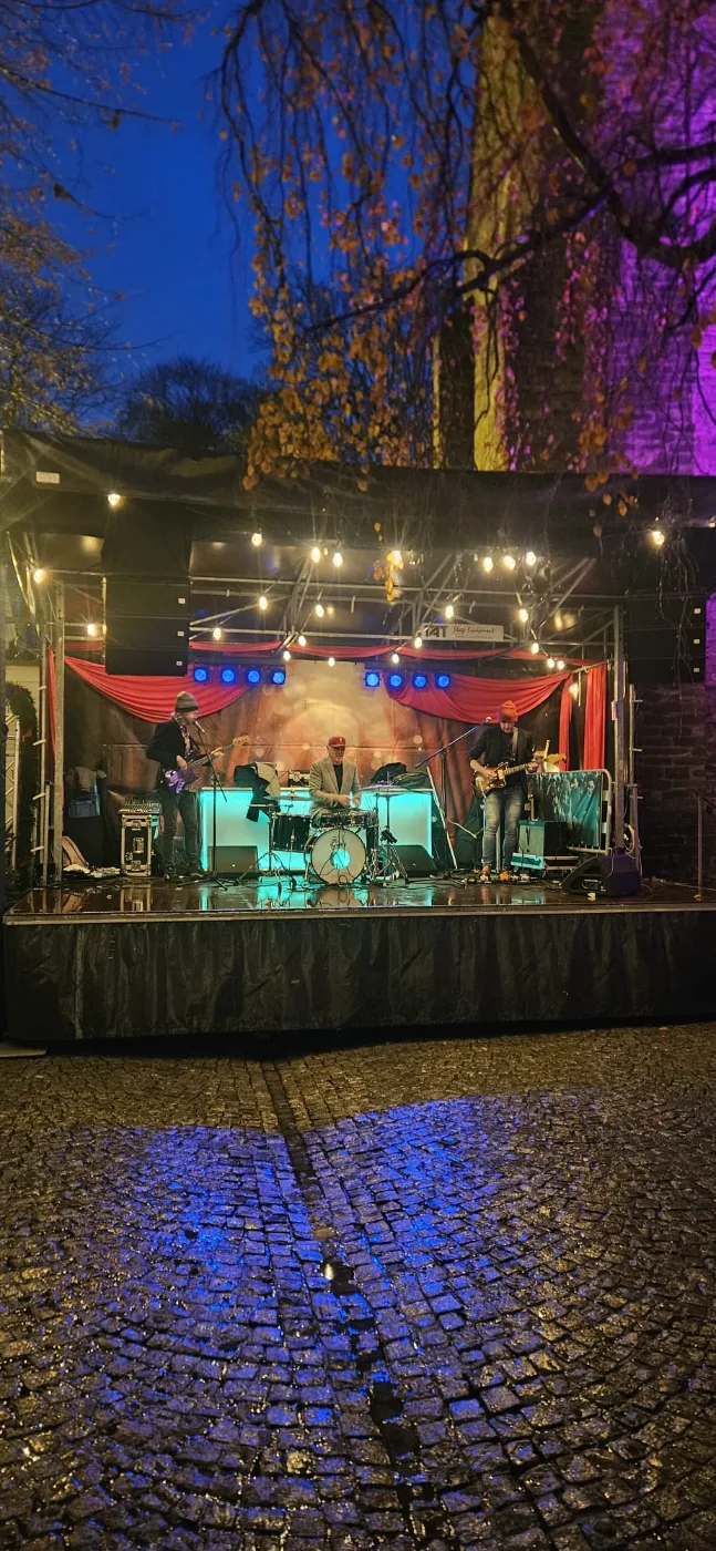 Band op een overdekt podium met string lights op een nat kasseiplein bij avond